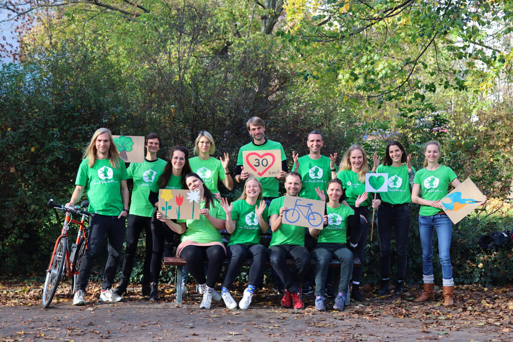 Gruppenbild vom Ökolöwen. Die Frauen und Männer tragen grüne T-Shirts mit dem Logo des Vereins, einem Löwen. Sie halten Schilder mit Blumen, Vögeln und weiteren Dingen, die Umweltschutz widerspiegeln, hoch. Im Hintergrund sind viele Bäume zu sehen.